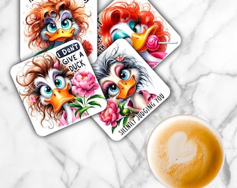 Puede incluir: Cuatro posavasos cuadrados con ilustraciones de patos de dibujos animados con pelo revuelto, ojos grandes y pestañas largas. El texto incluye It's fine it's okay, I don't give a duck y Silently judging you. Los diseños incorporan flores rosas y una piruleta.