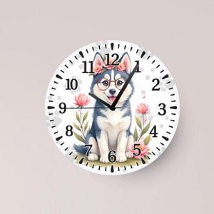 May include: Round wall clock featuring a watercolor illustration of a smiling husky dog wearing glasses and a flower crown. The clock face has black numbers and hands, with pink flowers and green leaves.