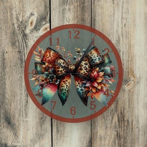 May include: A round wall clock with a decorative bow design. The bow features a leopard print pattern and is adorned with floral accents in shades of orange, red, and teal. The clock face is gray with red numerals and hands.
