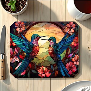 May include: A rectangular cutting board featuring a stained-glass design of two hummingbirds facing each other. The birds have blue, green, red, and orange feathers. The background is a sunset scene with flowers.
