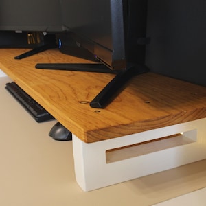 May include: A wooden monitor stand with a white base and a natural wood top. The stand is designed to elevate a computer monitor and create a more ergonomic workspace.