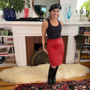 May include: A woman wearing a black tank top, a red leather pencil skirt, and black boots. She is also wearing a black beret. The background includes a fireplace, a mirror, and bookshelves.