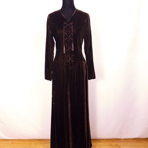 May include: A long, dark brown velvet dress with long sleeves and a V-neck. The bodice features a laced-up design with tassels. The dress is displayed on a black mannequin against a white backdrop and wooden floor.