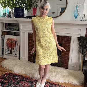 May include: A sleeveless, knee-length yellow dress with an embroidered floral pattern. The dress has a high neckline and is paired with white flats. The background includes a fireplace, a rug, and a mirror.