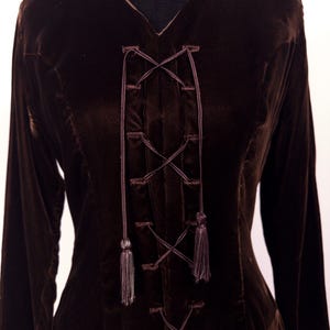 May include: A dark brown velvet dress with long sleeves and a V-neck. The bodice features a criss-cross lace-up design with tassels. The dress is displayed on a black mannequin.