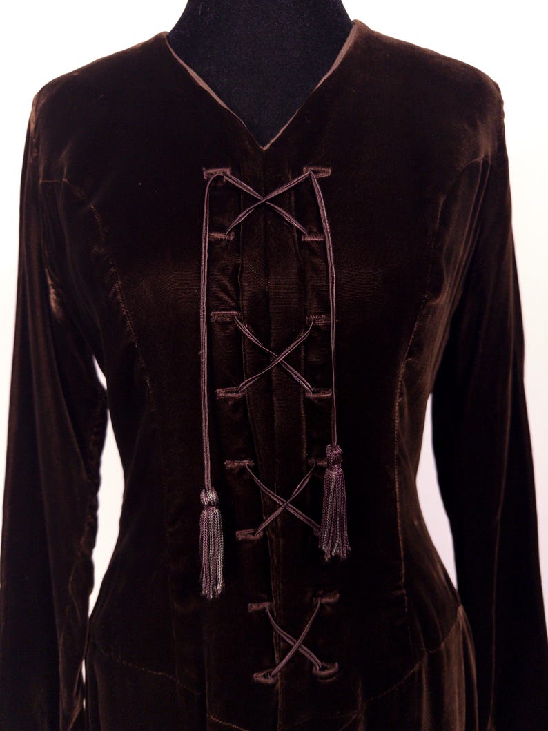 May include: A dark brown velvet dress with long sleeves and a V-neck. The bodice features a criss-cross lace-up design with tassels. The dress is displayed on a black mannequin.