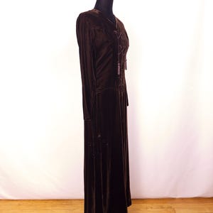 May include: A long, dark brown velvet dress with long sleeves and a V-neck. The dress features a lace-up detail at the bodice and is displayed on a black dress form. The dress has a floor-length hemline and a flowing silhouette.