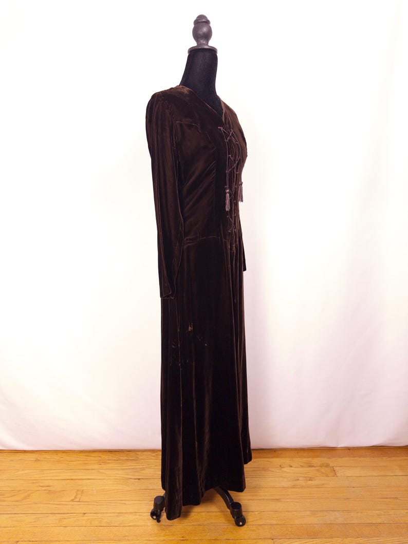 May include: A long, dark brown velvet dress with long sleeves and a V-neck. The dress features a lace-up detail at the bodice and is displayed on a black dress form. The dress has a floor-length hemline and a flowing silhouette.
