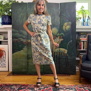 May include: A knee-length dress featuring a cream base with a repeating floral pattern in shades of blue and green. The garment has short sleeves, a round neckline with a dark trim, and a matching fabric tie belt at the waist.