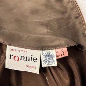 May include: Close-up of a vintage brown dress with a label that reads "Dress-ups by Ronnie Fashions." The label also includes the ILGWU union label and a size 11 tag. The dress is made of a shiny, silky material.