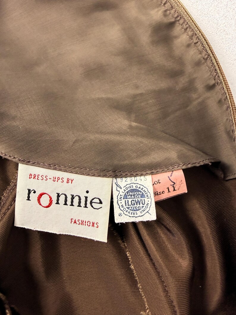 May include: Close-up of a vintage brown dress with a label that reads "Dress-ups by Ronnie Fashions." The label also includes the ILGWU union label and a size 11 tag. The dress is made of a shiny, silky material.