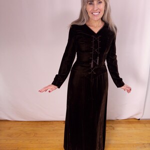 May include: A long, dark brown velvet dress with long sleeves and a V-neckline. The bodice features a decorative lace-up design with tassels. The dress has a fitted waist and a flowing skirt. The model is wearing silver shoes.