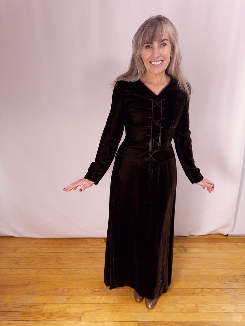 May include: A long, dark brown velvet dress with long sleeves and a V-neckline. The bodice features a decorative lace-up design with tassels. The dress has a fitted waist and a flowing skirt. The model is wearing silver shoes.