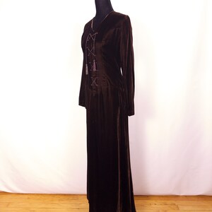May include: A long, dark brown velvet dress with long sleeves and a V-neckline. The bodice features a lace-up design with decorative tassels. The dress is displayed on a black mannequin against a white backdrop and wooden floor.