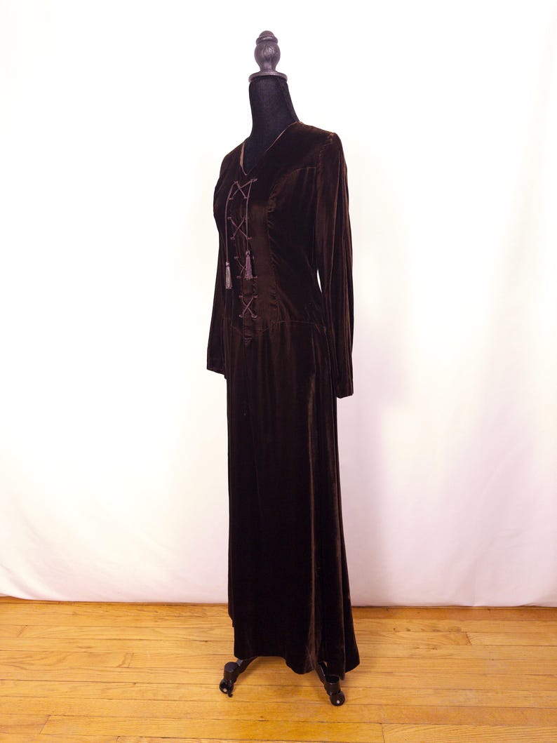 May include: A long, dark brown velvet dress with long sleeves and a V-neckline. The bodice features a lace-up design with decorative tassels. The dress is displayed on a black mannequin against a white backdrop and wooden floor.