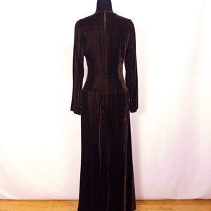 May include: A long, dark brown velvet dress with long sleeves and a high neckline. The dress is displayed on a black dress form against a white backdrop. The dress has a fitted bodice and a flowing skirt.