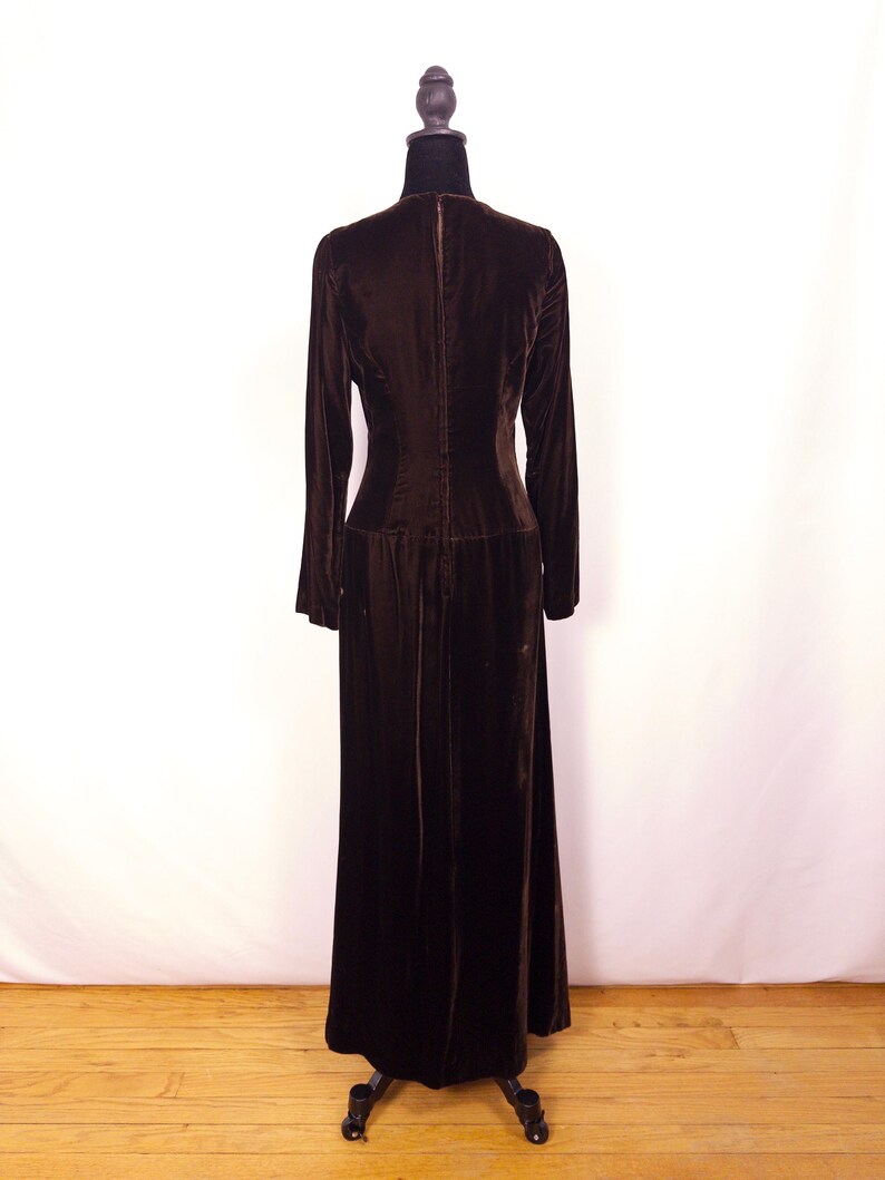 May include: A long, dark brown velvet dress with long sleeves and a high neckline. The dress is displayed on a black dress form against a white backdrop. The dress has a fitted bodice and a flowing skirt.