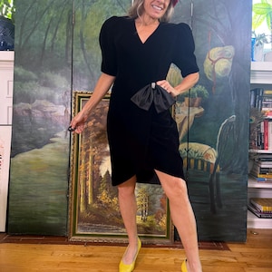 May include: A black velvet dress with short puff sleeves, a V-neckline, and a large black satin bow at the waist featuring a rhinestone accent. The dress has a wrap-style skirt. Also included is a multi-colored textured hat and yellow pointed-toe heels.
