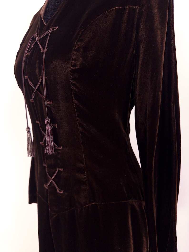 May include: A dark brown velvet top with long sleeves and a lace-up front. The top features a V-neckline and decorative tassels at the end of the lacing. The fabric has a soft, luxurious appearance.