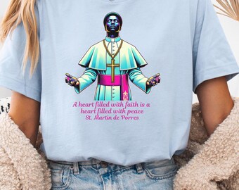 May include: A light blue t-shirt with a graphic of a person wearing a bishop's robe and a quote that reads "A heart filled with faith is a heart filled with peace. St. Martin de Porres."