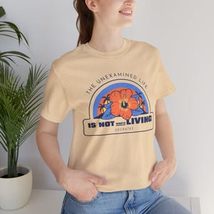 May include: A light yellow t-shirt with a graphic print of a hibiscus flower and the text "The unexamined life is not worth living - Socrates".