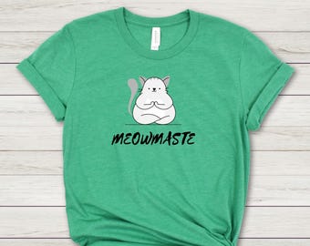 Peut inclure: T-shirt vert avec un chat de dessin animé en position de yoga. Le chat est blanc avec des accents gris et une queue grise. Le texte "MEOWMASTE" est imprimé en noir en dessous.