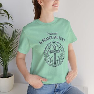 May include: A light green t-shirt with a graphic of a cross surrounded by flowers. The text "Centered in Prayer and Peace" is printed on the shirt.