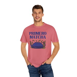 May include: A coral-colored t-shirt with the text "PRIMERO MATCHA" in blue above a blue teapot illustration with green leaves. The shirt is made of a soft material and is suitable for casual wear.