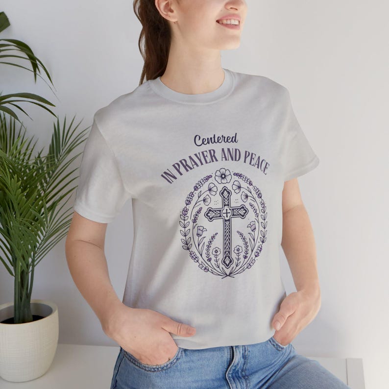 May include: A gray t-shirt with a floral design and a cross in the center. The text "Centered in Prayer and Peace" is printed on the shirt.