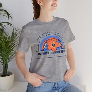 May include: Gray t-shirt with a graphic print of a hibiscus flower and palm trees. The text "The unexamined life is not worth living - Socrates" is printed on the shirt.