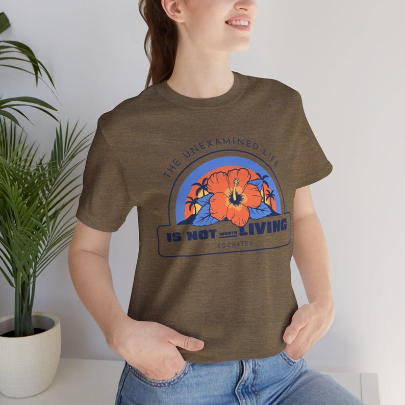 May include: A brown t-shirt with a graphic print of a hibiscus flower and palm trees. The text "The unexamined life is not worth living - Socrates" is printed on the shirt.