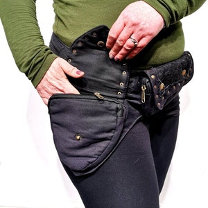 May include: A black canvas belt with multiple pockets and a flap with decorative studs. The belt has a zippered pouch and a smaller pouch with a flap. The belt is worn around the waist of a person.