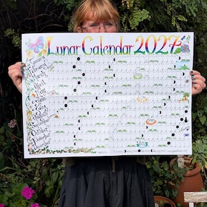May include: A large rectangular wall calendar titled Lunar Calendar 2027 with colourful hand-drawn lettering. The grid displays months from January to December, featuring moon phase icons, small illustrations, and seasonal labels across the white paper.
