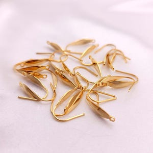 May include: A collection of gold-colored earring hooks, each featuring a leaf-shaped design. The hooks are scattered on a white, slightly textured surface. The hooks are made of a shiny, metallic material.