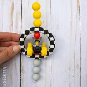 K&ouml;nnte beinhalten: Handgefertigter Autoschmuck mit einem kleinen gelb-schwarzen Spielzeugtraktor in einem schwarz-wei&szlig; karierten Ring. Der Schmuck hat gelbe und graue Perlen und ein wei&szlig;es Band zum Aufh&auml;ngen.