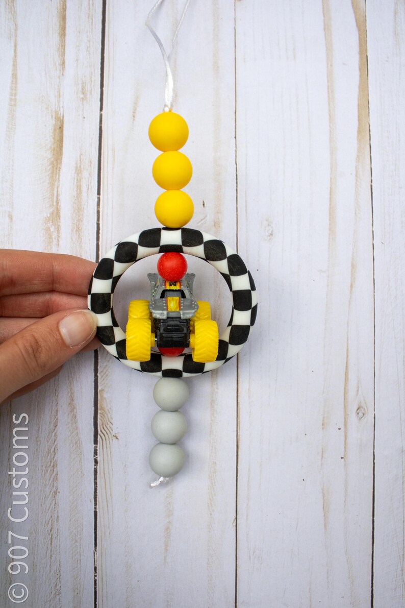 K&ouml;nnte beinhalten: Handgefertigter Autoschmuck mit einem kleinen gelb-schwarzen Spielzeugtraktor in einem schwarz-wei&szlig; karierten Ring. Der Schmuck hat gelbe und graue Perlen und ein wei&szlig;es Band zum Aufh&auml;ngen.