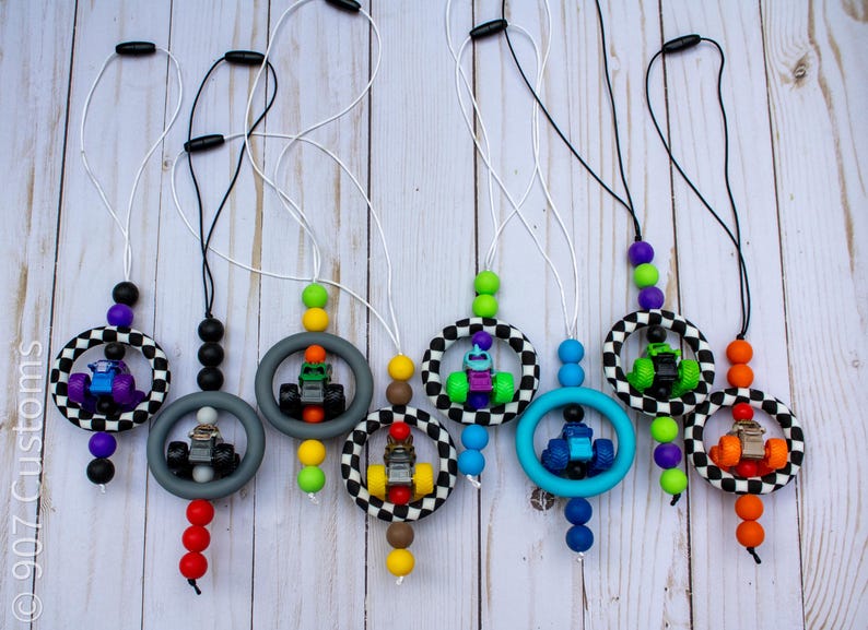 May include: A selection of car-themed necklaces. Each necklace has a small toy race car within a ring, with various bead colours and a black and white chequered ring. The necklaces are on white cords with black clasps.