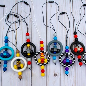 K&ouml;nnte beinhalten: Eine Sammlung von Schnullerhaltern f&uuml;r Babys mit Rennwagen-Designs. Jeder Halter hat eine farbenfrohe Perlenkette, einen Silikonring und ein kleines Spielzeug-Rennauto. Die Halter sind in verschiedenen Farben erh&auml;ltlich.