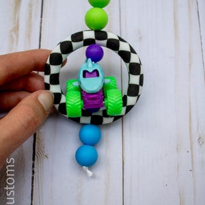 May include: A baby toy with a race car theme. The toy features a small, blue and purple monster truck inside a black and white chequered ring. Green and blue beads are attached to the toy with a white string.