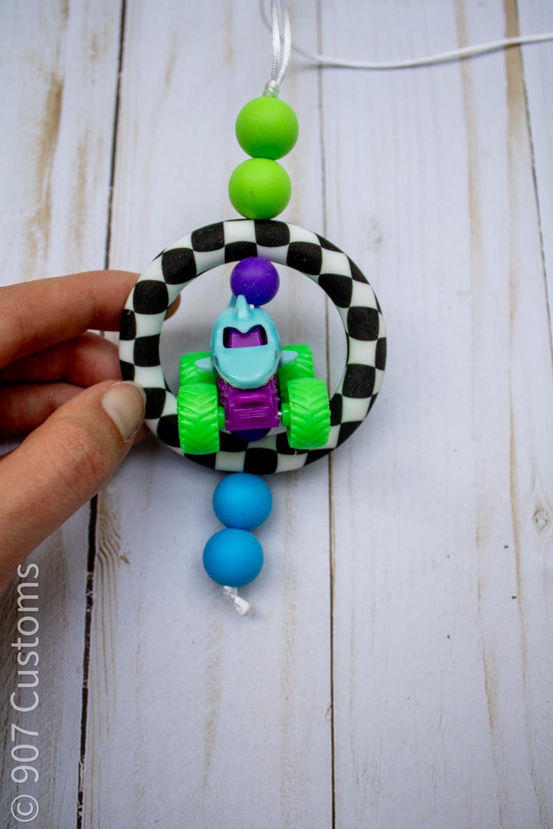 May include: A baby toy with a race car theme. The toy features a small, blue and purple monster truck inside a black and white chequered ring. Green and blue beads are attached to the toy with a white string.