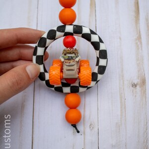 May include: A handmade car charm featuring a toy monster truck inside a black and white chequered ring. The charm has orange beads and a black cord for hanging. The toy truck has orange wheels and a tan body.