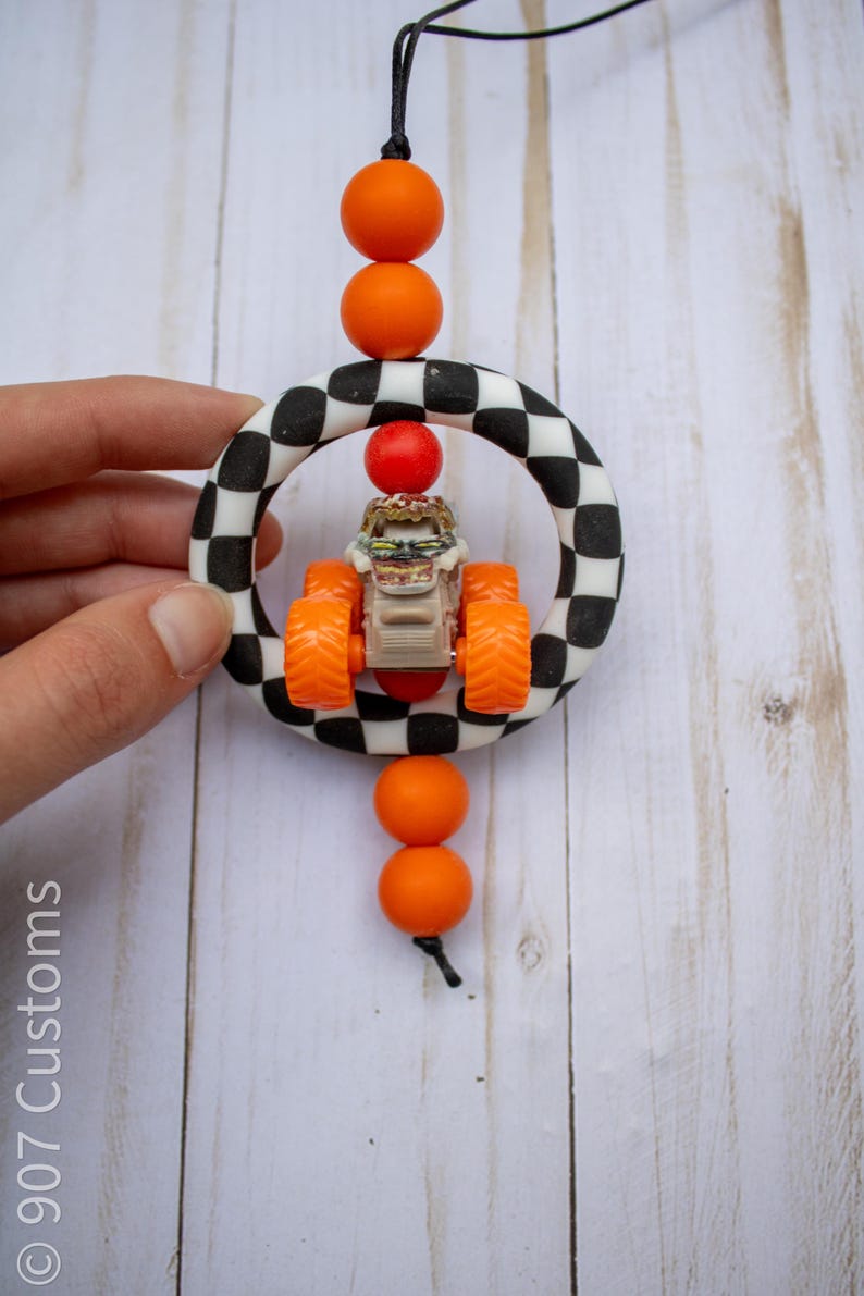 May include: A handmade car charm featuring a toy monster truck inside a black and white chequered ring. The charm has orange beads and a black cord for hanging. The toy truck has orange wheels and a tan body.