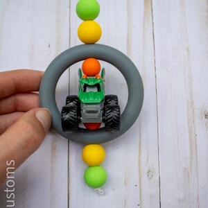 May include: A green and black toy monster truck is suspended within a grey ring, accented by colourful beads. The beads are yellow, green, and orange. A white cord is used to hang the toy. The text "907 Customs" is visible.