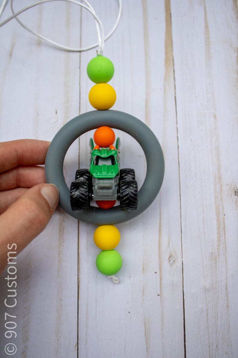 May include: A green and black toy monster truck is suspended within a grey ring, accented by colourful beads. The beads are yellow, green, and orange. A white cord is used to hang the toy. The text "907 Customs" is visible.