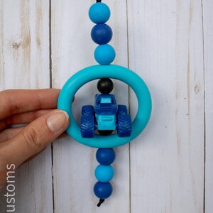 K&ouml;nnte beinhalten: Ein blau-t&uuml;rkisfarbenes Bei&szlig;spielzeug mit einem kleinen blauen Traktor in einem t&uuml;rkisfarbenen Ring. Das Spielzeug ist mit blauen Perlen in verschiedenen Farbt&ouml;nen und einer schwarzen Schnur aufgef&auml;delt. Der Hintergrund ist ein helles Holzmaserungsmuster.