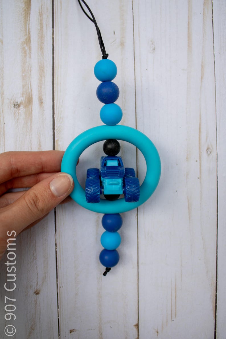 K&ouml;nnte beinhalten: Ein blau-t&uuml;rkisfarbenes Bei&szlig;spielzeug mit einem kleinen blauen Traktor in einem t&uuml;rkisfarbenen Ring. Das Spielzeug ist mit blauen Perlen in verschiedenen Farbt&ouml;nen und einer schwarzen Schnur aufgef&auml;delt. Der Hintergrund ist ein helles Holzmaserungsmuster.
