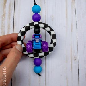 K&ouml;nnte beinhalten: Ein blaues und lila Monstertruck-Spielzeug ist in einem schwarz-wei&szlig; karierten Ring aufgeh&auml;ngt, der durch eine schwarze Schnur und farbige Perlen verbunden ist. Die Perlen sind blau, lila und schwarz. Der Spielzeug-Truck hat lila R&auml;der.