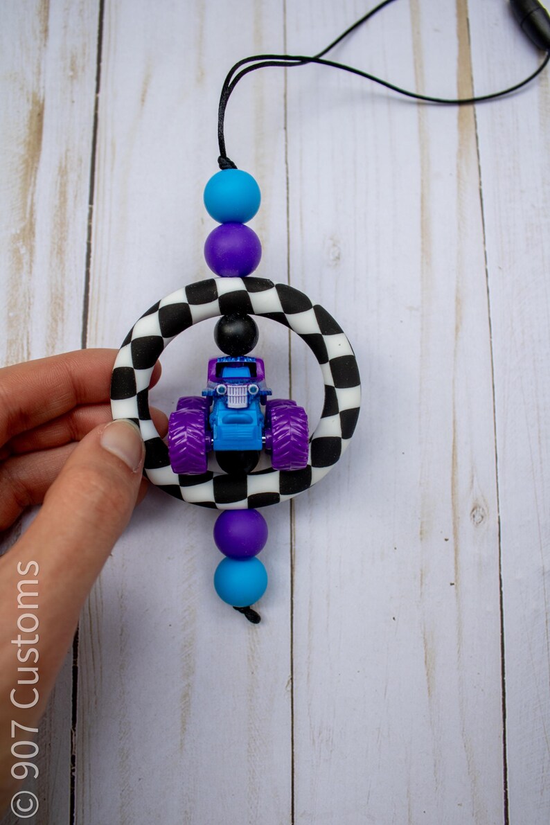 K&ouml;nnte beinhalten: Ein blaues und lila Monstertruck-Spielzeug ist in einem schwarz-wei&szlig; karierten Ring aufgeh&auml;ngt, der durch eine schwarze Schnur und farbige Perlen verbunden ist. Die Perlen sind blau, lila und schwarz. Der Spielzeug-Truck hat lila R&auml;der.