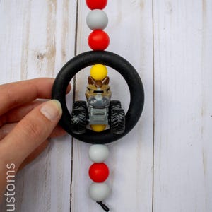 K&ouml;nnte beinhalten: Ein schwarzer Bei&szlig;ring mit einem Spielzeugtraktor im Inneren, aufgef&auml;delt mit roten, grauen und wei&szlig;en Perlen. Der Spielzeugtraktor ist braun, gelb und grau. Die Schnur ist schwarz.