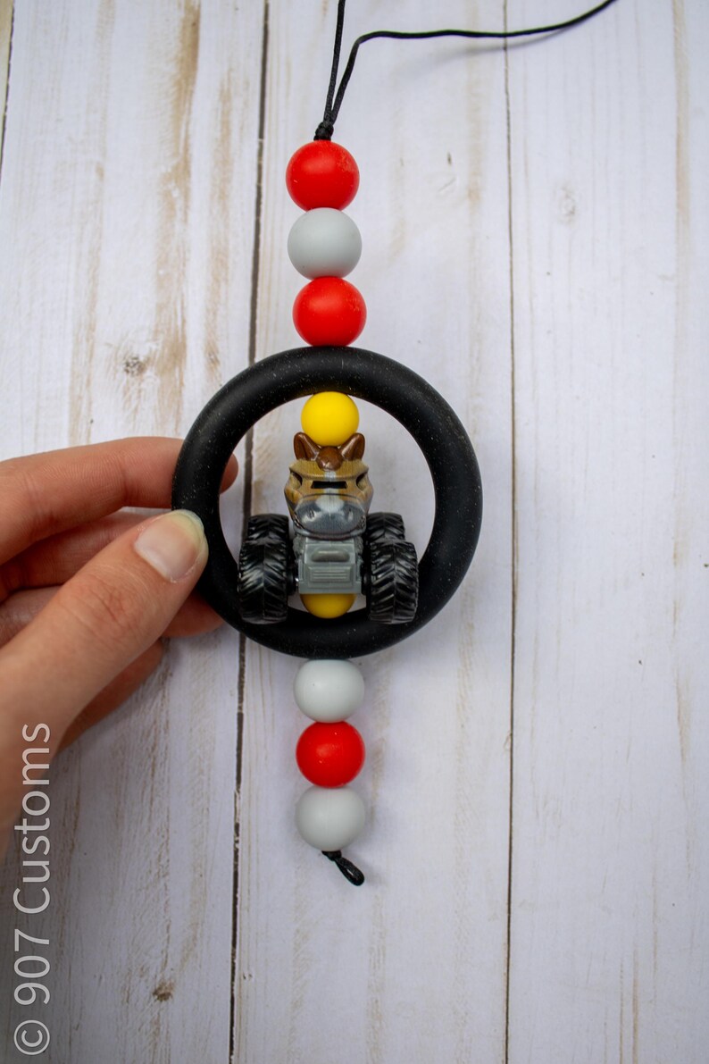 K&ouml;nnte beinhalten: Ein schwarzer Bei&szlig;ring mit einem Spielzeugtraktor im Inneren, aufgef&auml;delt mit roten, grauen und wei&szlig;en Perlen. Der Spielzeugtraktor ist braun, gelb und grau. Die Schnur ist schwarz.